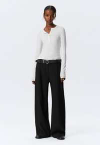 Wit lange mouwen henley-top met knoopdetails, gecombineerd met brede zwarte broeken en een zwarte riem, gedragen met zwarte platte schoenen.