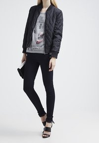 Chaqueta bomber negra acolchada sobre una camiseta gráfica, combinada con jeans ajustados negros y sandalias de tacón alto con tiras. Sostenido un clutch negro.