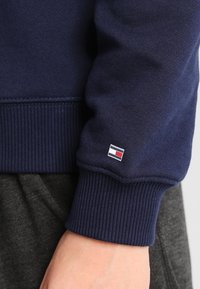 Felpa blu navy con polsini a coste, caratterizzata da un piccolo logo rettangolare in rosso, bianco e blu sulla manica. Texture morbida.