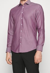 Muž v úzké, dlouhé košili s krátkým rukávem, která má bílé knoflíky a černé kalhoty, stojí na jednobarevném pozadí.