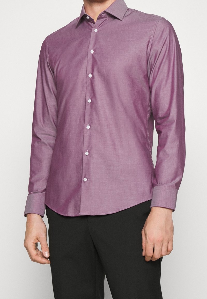 Muž v úzké, dlouhé košili s krátkým rukávem, která má bílé knoflíky a černé kalhoty, stojí na jednobarevném pozadí.