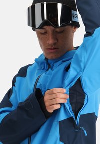 Blaue wasserdichte Jacke mit kontrastierenden dunkelblauen Akzenten, einer Reißverschlusstasche auf der Brust und einem Mesh-Futter. Das Model passt die innere Öffnung der Jacke an.