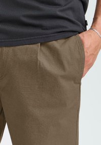 Persona vistiendo una camisa oscura y pantalones marrones, con una mano en el bolsillo, mostrando un detalle de un atuendo casual contra un fondo claro.