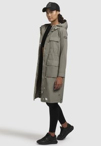 Parka de lungime mare, de culoare khaki, cu glugă, închidere cu fermoar și capse, multiple buzunare frontale și o etichetă de marcă pe mâneca stângă.