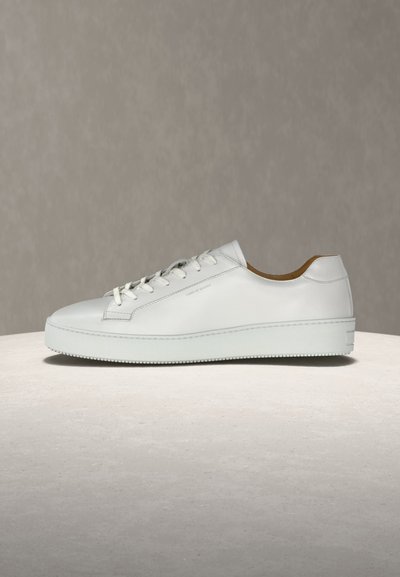 Baltas žemo profilio odinis sportbačis su baltomis raištelėmis ir storu padu, pastatytas ant šviesios dangos su neutraliu fonu.