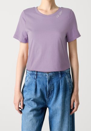 Vaaleanlilainen puuvillat-paita, jossa on pyöreä kaula-aukko ja lyhyet hihat, ja johon on brodeerattu pieni logo olkapään lähelle. Yhdistetty sinisiin farkkuihin.