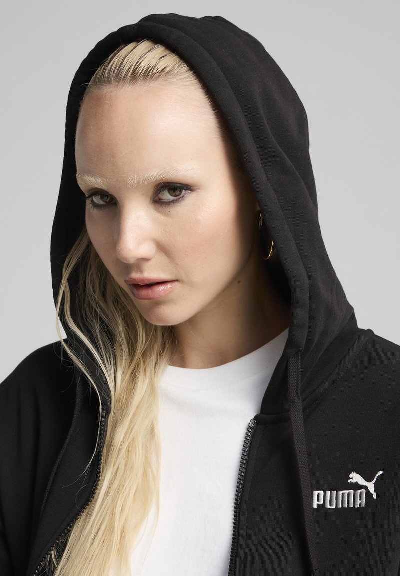 Sweat à capuche noir zippé avec cordon de serrage, arborant un logo PUMA brodé blanc sur le côté gauche de la poitrine, porté par-dessus une chemise blanche.