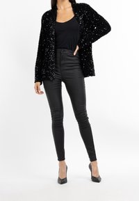 Cardigan nero con paillettes sopra una canotta nera, abbinato a jeans skinny neri a vita alta e tacchi a punta neri.