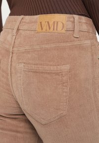 Κοντινό πλάνο στην πίσω τσέπη ανοιχτόχρωμων καφέ παντελονιών από κοτλέ με δερμάτινη ετικέτα μάρκας "VMD" στη μέση, συνδυασμένα με σκούρο καφέ ριμπ τοπ.