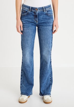 Persoon draagt blauwe jeans met rechte pijpen en witte sneakers, staand tegen een effen lichte achtergrond.