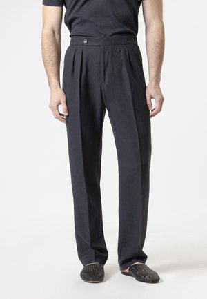 Hombre con pantalones plisados negros de talle alto y zapatos negros tejidos tipo slip-on, de pie frente a un fondo claro y liso.