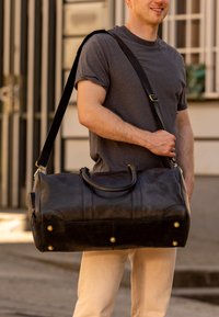 Sort læder duffel taske med guld hardware detaljer, dobbelte håndtag og en aftagelig skulderrem. Struktureret overflade med synlige sømme.