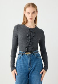 Grijze lange mouwen trui met gerimpeld voorpand, voorzien van knoopsluitingen en een gladde textuur. Gekleed met hoge taille blauwe spijkerbroek.