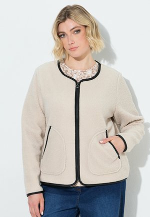 Kvinde med kort blond hår iført en beige fleecejakke med lynlås og sort kant samt blå jeans, med den ene hånd i lommen.