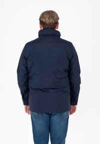 Giacca impermeabile blu navy con colletto alto, maniche lunghe e vestibilità rilassata. Il materiale appare liscio con una leggera lucentezza. I dettagli sono minimi.