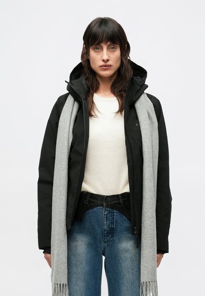 Giacca nera con cappuccio e cerniere, indossata sopra un maglione crema e abbinata a jeans in denim di lavaggio medio e una sciarpa grigio chiaro.