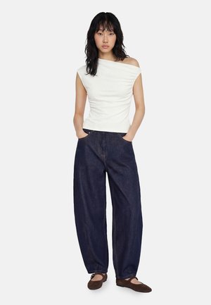 Mujer de pie con las manos en los bolsillos, vistiendo un top blanco sin hombros, vaqueros sueltos azul oscuro y zapatos planos marrones sobre fondo blanco.