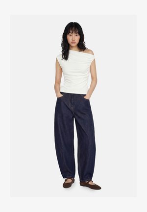 Mujer de pie con las manos en los bolsillos, vistiendo un top blanco sin hombros, vaqueros sueltos azul oscuro y zapatos planos marrones sobre fondo blanco.