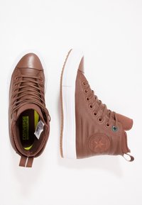 Hnědé vysoké tenisky vyrobené z hladké kůže, s bílou gumovou podrážkou, texturovanými akcenty a kulatým logem na boku.