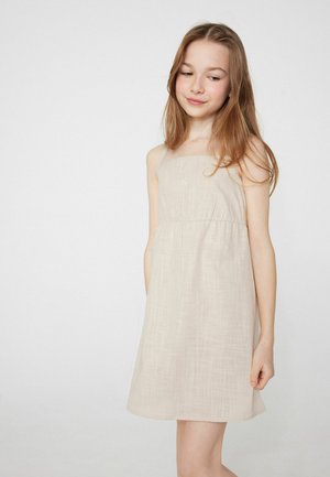 Mladá dívka s dlouhými vlasy v béžových šatech bez rukávů stojí před jednobarevným bílým pozadím a mírně se dívá dolů.