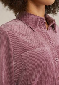 Kostymskjorta i mjuk mauve med vertikal ribbad textur, knappkrage, frampocket och matchande knappar.