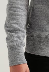 Grauer Pullover aus sweatshirtartigem Material mit strukturierter Oberfläche, gerippten Bündchen und einem kleinen Markenschild an der unteren Seite. Figurbetonter Schnitt mit langen Ärmeln.