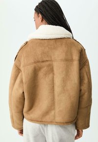 Donna con capelli intrecciati che indossa una giacca in suede beige con colletto in pile bianco, vista di spalle su uno sfondo bianco.