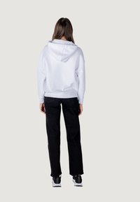Sudadera blanca con capucha ajustable, combinada con pantalones negros. La tela parece suave y el corte es relajado. Incluye un bolsillo tipo canguro.