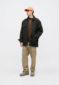 Chaqueta a cuadros en negro y gris con una capa interior marrón, pantalones cargo beige y botas de color verde olivo. Una gorra naranja completa el atuendo.