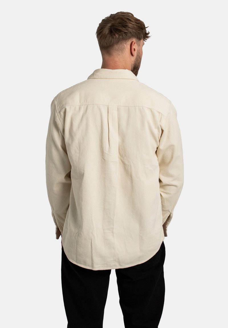 Beige corduroy shirt with a relaxed fit, featuring a back pleat and buttoned cuffs. Worn with black pants.