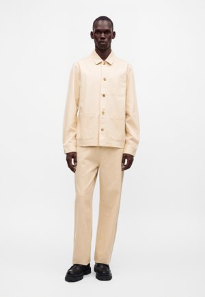 Homme debout portant une veste beige boutonnée et un pantalon assorti avec des chaussures noires sur un fond blanc uni.