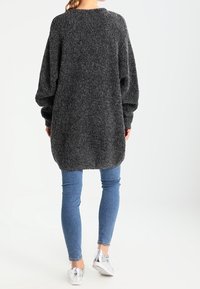 Överdimensionerad svart tröja med en texturerad, fluffig yta, försedd med långa ärmar och rundad nederkant, tillsammans med jeans och silverfärgade sneakers.