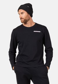 Schwarzes Langarmshirt mit rundem Ausschnitt, das ein pinkes "Rossignol"-Logo auf der Brust hat. Das Modell trägt eine schwarze Strickmütze.