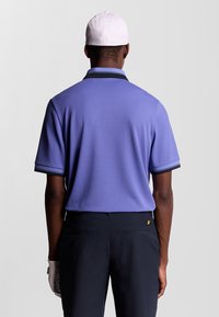 Uomo che indossa una polo a maniche corte viola, un cappellino bianco, pantaloni neri e un guanto bianco, in piedi rivolto di spalle su uno sfondo neutro.