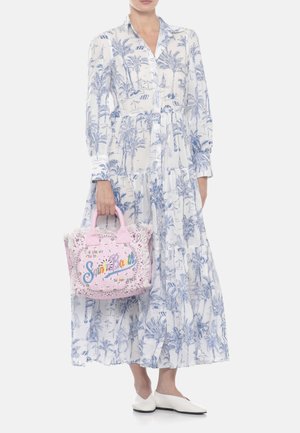 Donna con un abito lungo bianco e blu con stampa di palme che tiene una borsa rosa con scritta colorata "Saint Barth" e scarpe basse bianche.