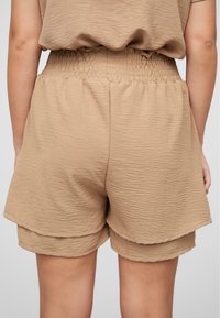 Shorts texturés en tan avec une taille froncée et une coupe décontractée. Le tissu semble léger et légèrement froissé pour une dimension supplémentaire.