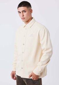 Camisa de botones color crema con textura acanalada, que presenta botones a presión plateados y un sutil logotipo bordado en el pecho.
