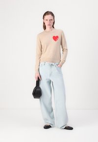 Beżowy sweter wełniany z czerwoną łatką w kształcie serca, w połączeniu z jasnoniebieskimi szerokimi spodniami dżinsowymi i czarnymi balerinami, uzupełniony małą czarną torebką.