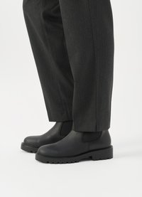 Melni ādas potīšu zābaki ar elastīgām sānu paneļiem un biezu, izturīgu zoli, apvienoti ar pelēkām, tekstūrētām sašaurinātām biksēm.