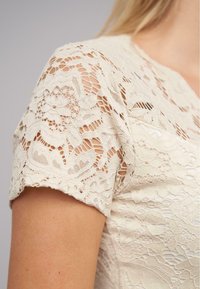 Lace top met korte mouwen in crèmekleur, met een bloemenpatroon en open weefdetails, die een ingewikkelde textuur en ontwerp laat zien.