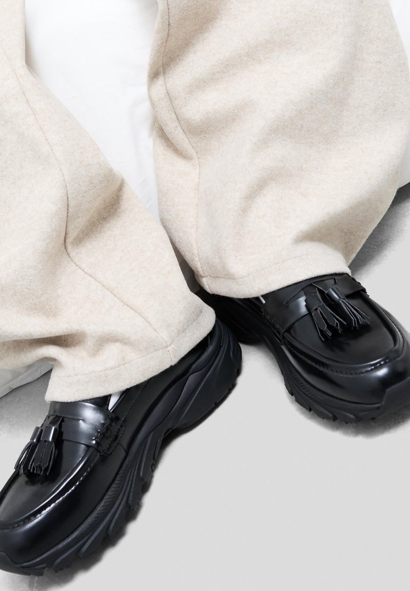 Čierne kožené loafers s pampúšikmi, s hrubou, textúrovanou podrážkou, v spojení s svetlo béžovými, mäkkými, mierne voľnými nohavicami.