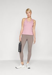 Canottiera sportiva rosa chiaro, leggings beige, tiene in mano una borraccia nera. L'outfit include scarpe da ginnastica bianche con accenti neri. Design semplice.