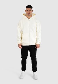 Crème zip-hoodie med stor huva och framfickor, tillsammans med svarta joggare och vita sneakers mot en enhetlig bakgrund.