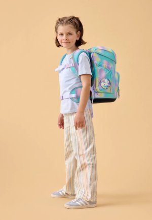 Un sac à dos aux couleurs pastel, avec des motifs circulaires, des bretelles réglables et une poche avant ; porté avec une chemise bleu clair et un pantalon rayé.