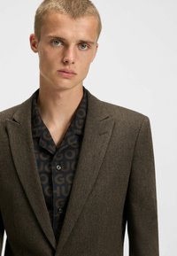 Brauner Fischgrät-Blazer mit Reverskragen über einem dunklen Hemd mit großem Logomuster. Glatte Stofftextur mit strukturiertem Schnitt.