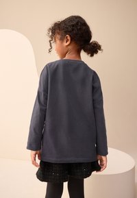 Temno siva dolga majica z dolgimi rokavi in okroglim izrezom, sproščenega kroja, prikazana od zadaj. Oblečena s črno teksturirano krilo in nogavicami.