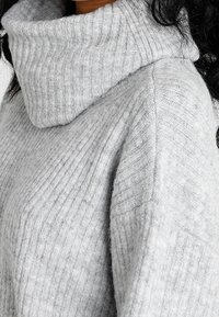 Vaaleanharmaa ribbikuvioinen pooloneule, jossa on pehmeä rakenne. Neuleessa on leveä, taitettu kaulus ja hieman laskeutuvat olkapäät, jotka tarjoavat rentoa istuvuutta.