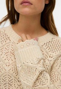 Femme portant un pull beige en tricot avec des poignets en dentelle, tenant le col de la main droite, montrant les détails du tissu et de la texture.