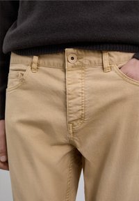 Beige bomuldsbukser med glat tekstur, der har lynlåslukning, knaplukning og forlommer; båret med en mørk trøje.