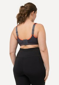 Sujetador deportivo en gris con acentos naranjas, tirantes ajustables y cierre posterior con gancho y ojal. Combinado con leggings negros de cintura alta.